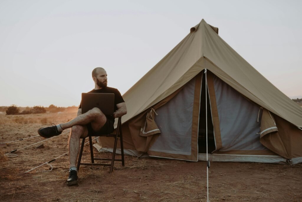 Bearded man working on a laptop outside a tent in a serene campsite setting, embracing remote work in nature.